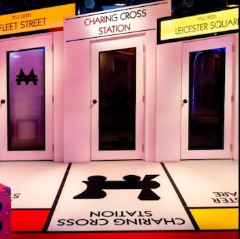 Tres puertas blancas con paneles oscuros, letreros de "Fleet Street", "Charing Cross Station" y "Leicester Square", y secciones del tablero de Monopoly en el suelo