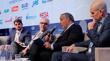 Cuatro hombres sentados en un panel de discusión; tres llevan traje y uno camisa con saco. El hombre central sostiene un micrófono. En primer plano hay botellas de agua y un vaso, con logos de patrocinadores al fondo