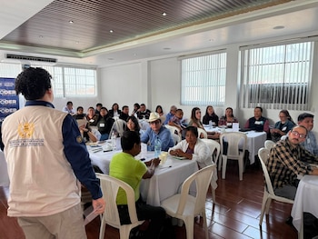 Sala con varias mesas blancas, sillas, un grupo de personas sentadas y un orador de espaldas con chaleco con logo