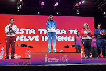 Cinco mujeres de pie sobre un escenario con podio, medallas y vestimenta deportiva frente a una gran pantalla roja con iluminación superior