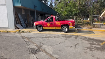 Una camioneta roja de Bomberos UNAM con logotipos amarillos está estacionada en el pavimento de un campus universitario, con edificios y árboles al fondo