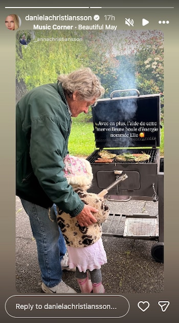Un hombre mayor con chaqueta verde y una niña con abrigo estampado junto a una barbacoa humeante con comida en el exterior