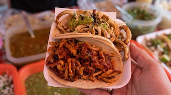 Mano que sostiene un plato con dos tacos rellenos de tripa de pollo sobre tortillas de maíz, con recipientes de salsa al fondo.