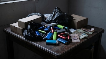 Varios vapeadores de colores llamativos y billetes mexicanos están apilados desordenadamente sobre una mesa de madera oscura, junto a cajas de cartón y bolsas negras.