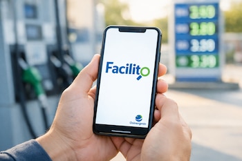 Manos sostienen un smartphone con la app Facilito (logo azul y verde) y Osinergmin en pantalla. Al fondo, surtidores de combustible y cartel de precios de estación.