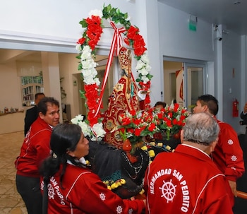 Grupo de personas en chaquetas rojas lleva una estatua de San Pedro, decorada con flores rojas y blancas, en una procesión interior