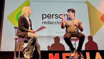 Una mujer con hojas y un hombre con micrófono hablan sentados en banquetas sobre un escenario frente a una pantalla