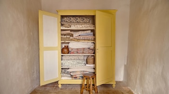 Armario de madera amarillo con puertas abiertas lleno de textiles doblados en estantes, con un jarrón y una cesta. Un taburete de madera se sitúa delante. Paredes texturizadas.
