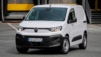 Vista frontal tres cuartos de una furgoneta comercial Citroën Berlingo blanca estacionada sobre asfalto gris, con un edificio amarillo y negro al fondo