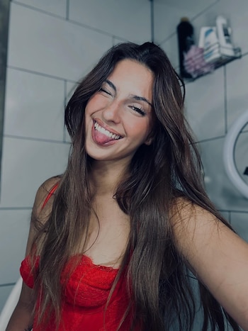 Primer plano de Lady M sonriendo y sacando la lengua, con cabello castaño largo y un top rojo, frente a un fondo de azulejos claros