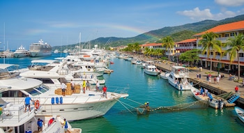 Múltiples yates, botes pequeños, dos cruceros grandes, edificios, palmeras, montañas, muelle con personas caminando y un arcoíris en el cielo.