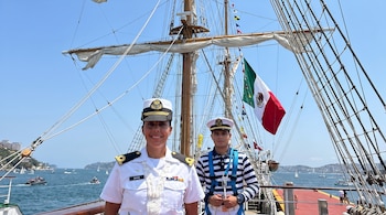Dos personas con uniformes navales posan en la cubierta de un buque de vela con mástiles y jarcia, una bandera de México y el mar con otras embarcaciones.