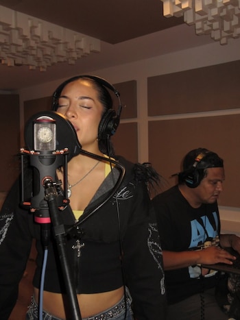Una mujer con auriculares canta frente a un micrófono en un estudio de grabación, sus ojos cerrados. Un hombre con auriculares opera un equipo en el fondo