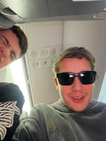 Primer plano de dos hombres jóvenes en el interior de un avión. Un hombre con gafas de sol oscuras sonríe en primer plano. Otro hombre sonríe parcialmente visible