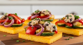 Tres bruschettas de polenta sobre una tabla de madera, con salteado de morrón rojo, zucchini, cebolla morada, champiñones y perejil picado.
