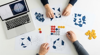 Mesa blanca con una laptop, cuya pantalla muestra un cerebro y gráficos. Varias hojas con patrones geométricos, fichas de colores y dos pares de manos.
