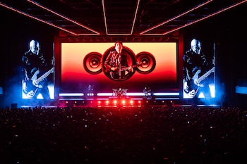 Escenario de concierto oscuro con pantallas gigantes que muestran a músicos de Soda Stereo tocando, rodeado de luces rojas y azules sobre una multitud