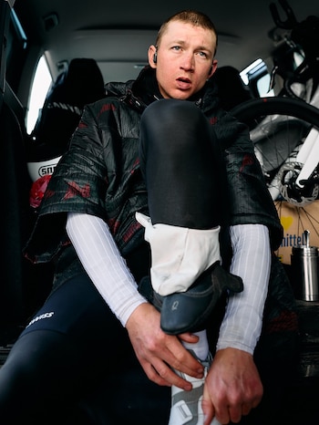 Hombre joven con chaqueta oscura y camiseta de manga larga blanca sentado dentro de la parte trasera de un coche, atándose un zapato, con una rueda de bicicleta visible