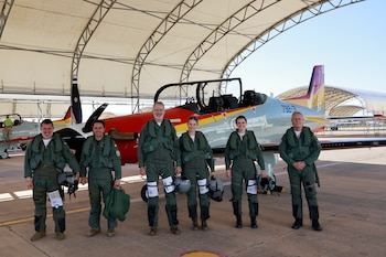Seis personas con trajes de vuelo verdes, chalecos y cascos posan frente a un avión militar rojo, gris y amarillo bajo la estructura de un hangar abierto