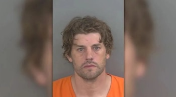 Primer plano de un hombre caucásico con cabello rizado y barba, vestido con un uniforme naranja, mirando directamente a la cámara para una foto policial