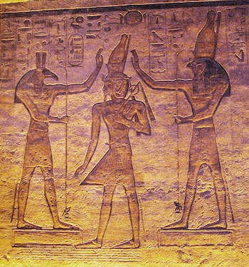 Un relieve de piedra con tres figuras de deidades egipcias, dos con cabezas de halcón y la central con corona, junto a jeroglíficos tallados