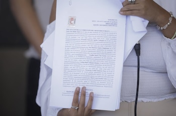 Una persona sostiene un documento oficial de la Fiscalía contra la Corrupción del Ministerio Público con texto impreso, lleva un anillo y una pulsera. Hay un cable negro