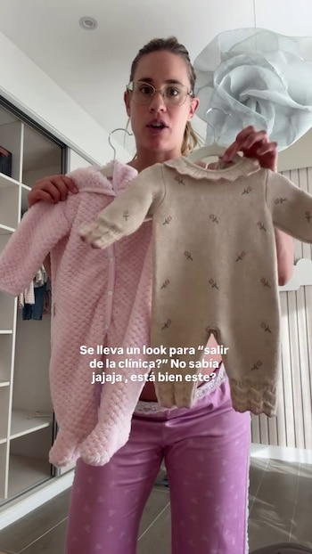 Una mujer con gafas sostiene dos atuendos de bebé en perchas: uno rosa afelpado y otro beige tejido con diseños florales. Al fondo, un armario abierto