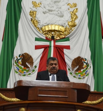 Un hombre con bigote, traje oscuro y corbata gris claro está de pie detrás de un podio de madera. Detrás de él, una bandera de México drapeada con un escudo