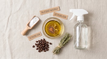 Un tazón con mezcla de aceite, una pala con bicarbonato, un pulverizador, granos de café, romero y etiquetas sobre una tela de lino.