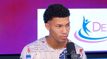 Un hombre joven de cabello rizado viste una camiseta estampada y se encuentra ante un micrófono en un estudio.