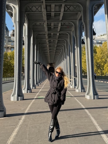 Una mujer con abrigo negro, botas y gafas de sol posa con el brazo levantado bajo las columnas de metal de un puente