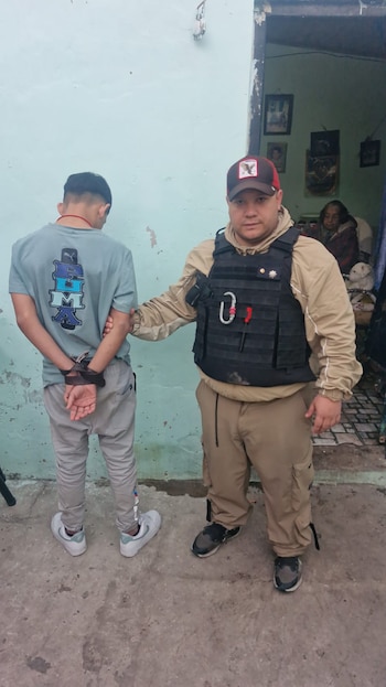 Un agente con chaleco táctico sujeta a un joven esposado por la espalda, de pie frente a una pared azul claro, con una mujer sentada visible al fondo