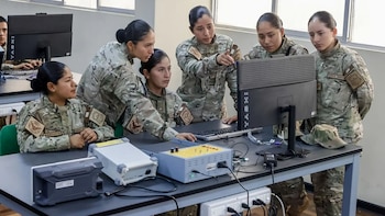 Seis mujeres con uniforme militar camuflado observan la pantalla de una computadora sobre una mesa con equipos electrónicos