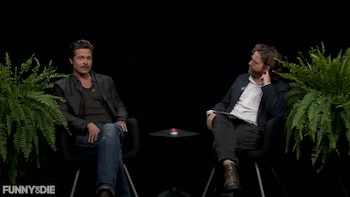 Brad Pitt y Zach Galifianakis sentados en sillas negras con un fondo oscuro y helechos a cada lado, un botón rojo en una mesa central