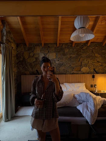 Mujer se toma una selfie en un espejo de una habitación de hotel, sujeta un mate; se ven una cama, una pared de piedra, techo de madera y una lámpara