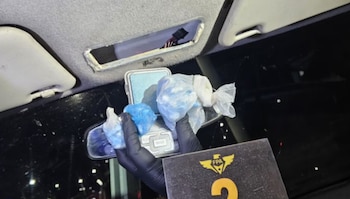 Una mano enguantada de negro sostiene bolsas con sustancias blancas y azules sobre una balanza digital dentro del habitáculo de un auto, con el techo parcialmente desmantelado