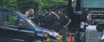 Oficiales de la oficina del sheriff y vehículos policiales negros estacionados frente a un fondo de vegetación, con cinta roja visible en un automóvil