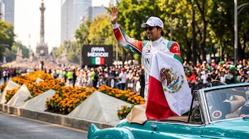 Un piloto con traje de carreras, gorra y bandera mexicana saluda de pie desde un automóvil descapotable en una avenida con público.