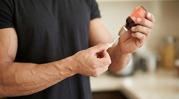 Primer plano de las manos de un joven musculoso con camiseta negra, extrayendo un líquido de un frasco rojo y ámbar con una jeringa.
