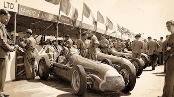 Ilustración en sepia de coches de carreras clásicos, pilotos y mecánicos en un pit lane, con carteles que nombran a Fangio, Farina y Fagioli, y banderas.