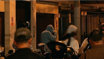 Un individuo con sudadera gris de capucha cubre su rostro frente a un edificio en una calle iluminada de noche