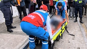 Dos paramédicos atienden a una persona con el rostro pixelado en una camilla con ruedas. Varias personas, algunas con uniforme, observan la escena