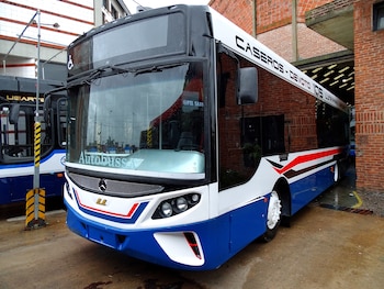Vista frontal de un autobús moderno de dos pisos, blanco y azul con franjas rojas y negras, estacionado junto a una pared de ladrillo