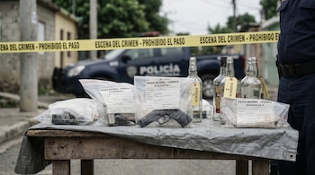 Mesa forense improvisada en República Dominicana con evidencias: una pistola en bolsa sellada y botellas de alcohol vacías, numeradas; cinta policial al fondo.