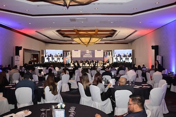 Panel de oradores en un escenario con pantallas, banderas de Guatemala y Estados Unidos, y un público asistente en mesas redondas