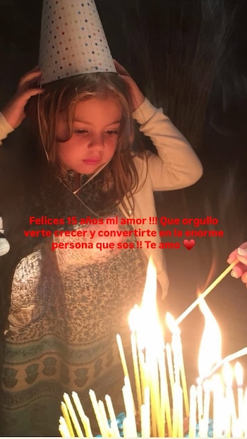 Una niña pequeña con un gorro cónico de fiesta observa las velas encendidas sobre un pastel mientras encienden otra vela