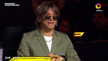Primer plano de Joaquín Levinton con gafas de sol, blazer verde, camiseta blanca y cadena dorada, sentado con fondo oscuro y luces naranjas