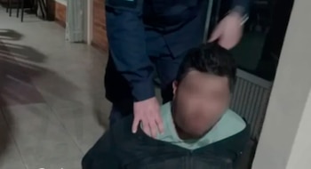 Hombre con camisa verde y chaqueta oscura, con rostro borroso, es sujetado por otra persona vestida de uniforme azul en el interior de una casa