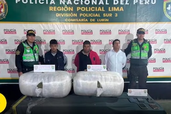 Dos policías y tres hombres se paran detrás de una mesa con dos fardos grandes de droga y celulares incautados. Banner de la Policía Nacional del Perú al fondo