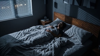 Una mujer está acostada en una cama con ropa de cama gris en una habitación a oscuras iluminada por franjas de luz